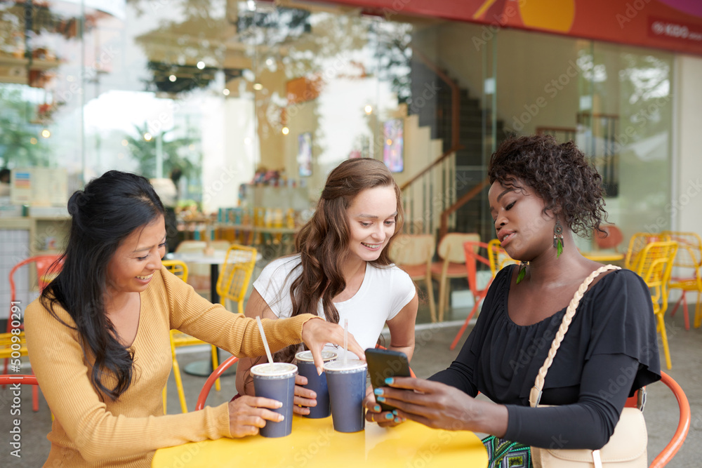 Young woman showing photos on smartphone to friends when they are having soft drinks at cafe table Young woman showing photos on smartphone to friends when they are having soft drinks at cafe table