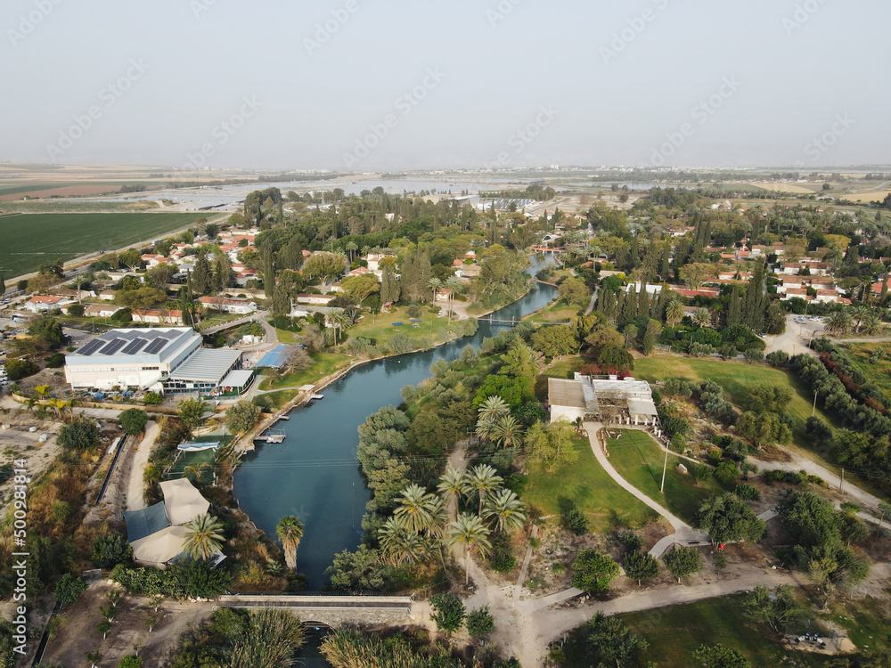Aerial panorama view of Kibbutz Nir David in Northern Israel with nahal Amal river turquoise water between houses and palm trees surrounding agricultural land.