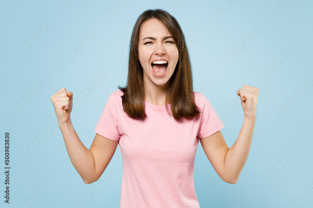 Young excited happy caucasian woman 20s in pink t-shirt doing winner gesture celebrate clenching fists say yes isolated on pastel plain light blue background studio portrait. People lifestyle concept. Young excited happy caucasian woman 20s in pink t-shirt doing winner gesture celebrate clenching fists say yes isolated on pastel plain light blue background studio portrait. People lifestyle concept.