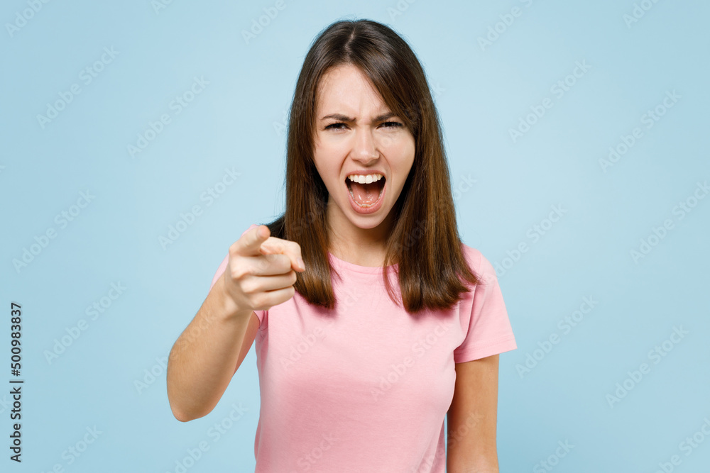 Fototapeta premium Young sad abuser angry caucasian woman 20s wearing pink t-shirt point index finger camera on you scream shout isolated on pastel plain light blue background studio portrait. People lifestyle concept.