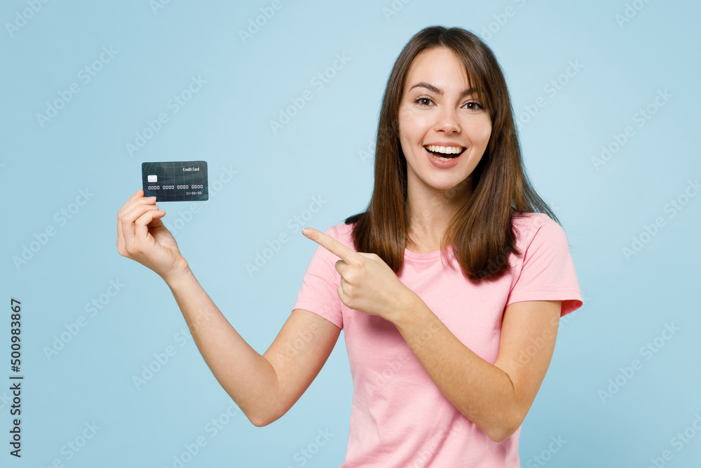 Young smiling happy cool fun woman 20s wearing pink t-shirt hold in hand point index finger on credit bank card isolated on pastel plain light blue background studio portrait People lifestyle concept Young smiling happy cool fun woman 20s wearing pink t-shirt hold in hand point index finger on credit bank card isolated on pastel plain light blue background studio portrait People lifestyle concept
