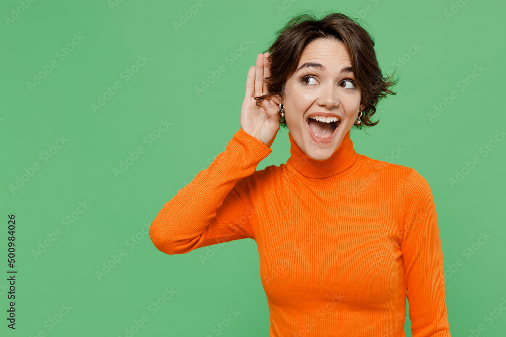 Young curious nosy woman 20s wear casual orange turtleneck try to hear you overhear listening intently isolated on plain pastel light green color background studio portrait. People lifestyle concept.