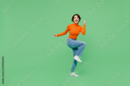 Foto Full body young woman 20s wear orange turtleneck doing winner gesture celebrate clenching fists say yes raise up leg isolated on plain pastel light green color background