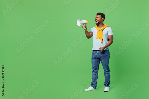 Full body young man of African American ethnicity 20s wear blue t-shirt hold scream in megaphone announces discounts sale Hurry up isolated on plain green background studio. People lifestyle concept.