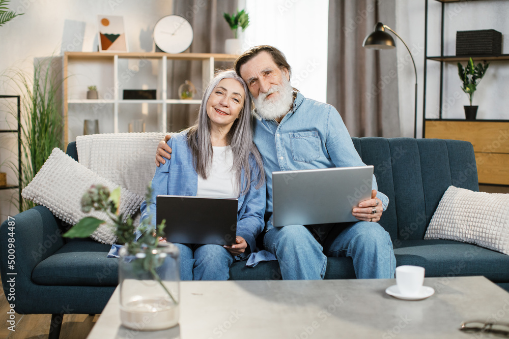 Quality time. Upbeat senior husband and wife holding a laptops in their laps and surfing the Internet together while sitting on the sofa in their living room. Quality time. Upbeat senior husband and wife holding a laptops in their laps and surfing the Internet together while sitting on the sofa in their living room.
