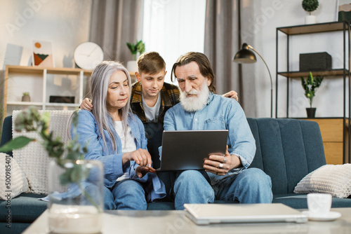 Family leisure, technology concept. Young grandson hugging his grandmother and grandfather in casual domestic clothes, sitting together on soft couch at home and watching interesting movie on laptop