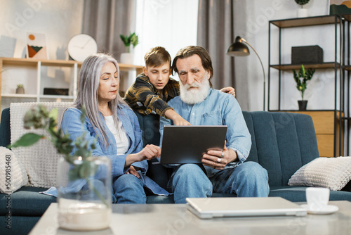 Pleasant happy smiling family of grandparents and grandson sitting on sofa at home and making online shopping together. Young happy boy shows something on the screen to his grandmother and grandfather
