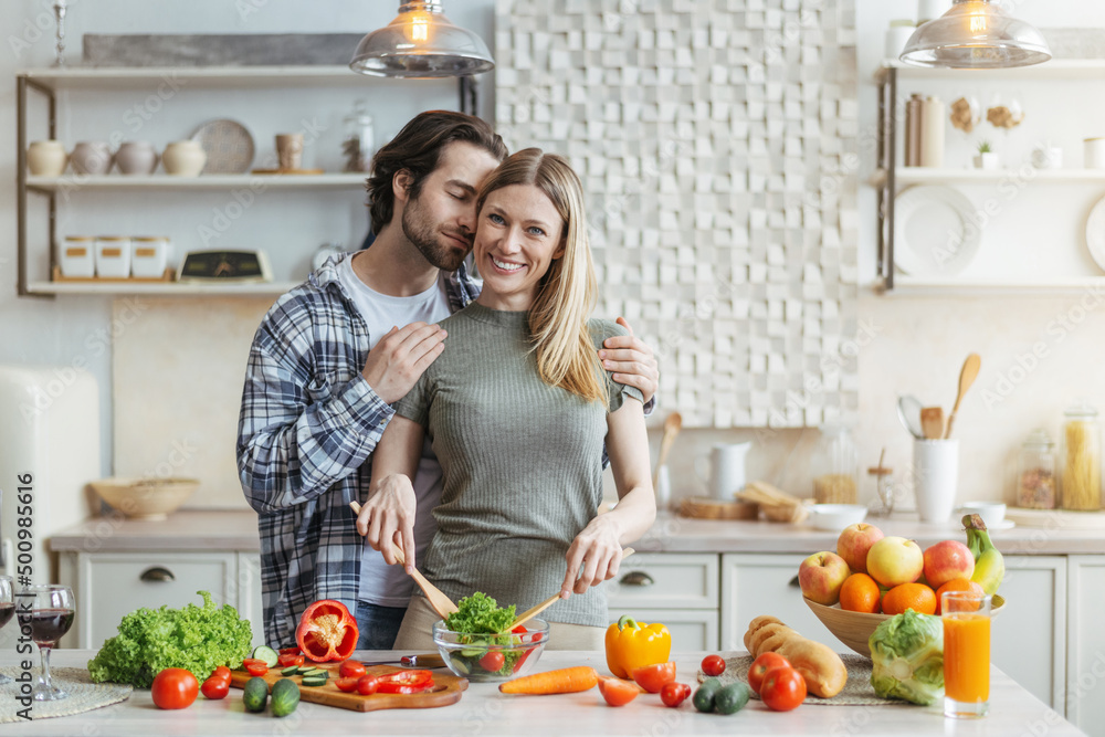 Smiling young european woman prepares salad, man with stubble hugs wife at table with organic vegetables Smiling young european woman prepares salad, man with stubble hugs wife at table with organic vegetables
