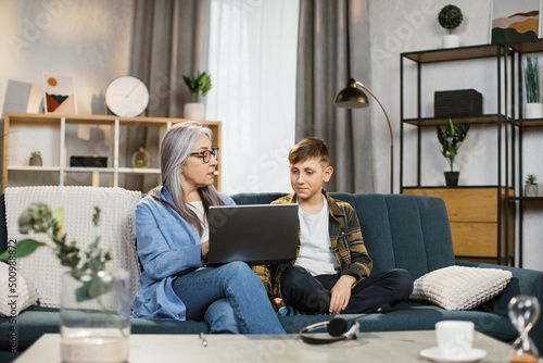 Young school age caucasianboy explaining his elderly retired mother how to use a laptop sitting on a blue sofa in a bright living room. Grandson teach senior elder to surf internet using laptop pc.
