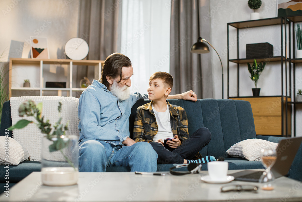 Happy grandfather with grey beard cuddling smiling small child boy playing online mobile game together at home. Joyful different generations family using funny smartphone applications, resting on sofa Happy grandfather with grey beard cuddling smiling small child boy playing online mobile game together at home. Joyful different generations family using funny smartphone applications, resting on sofa