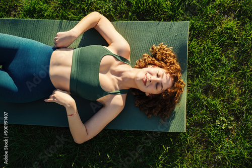 Top view of beautiful young happy sportive woman wearing green sports bra and blue yoga pant. Closed eyes and smiling while lying on yoga mat.
