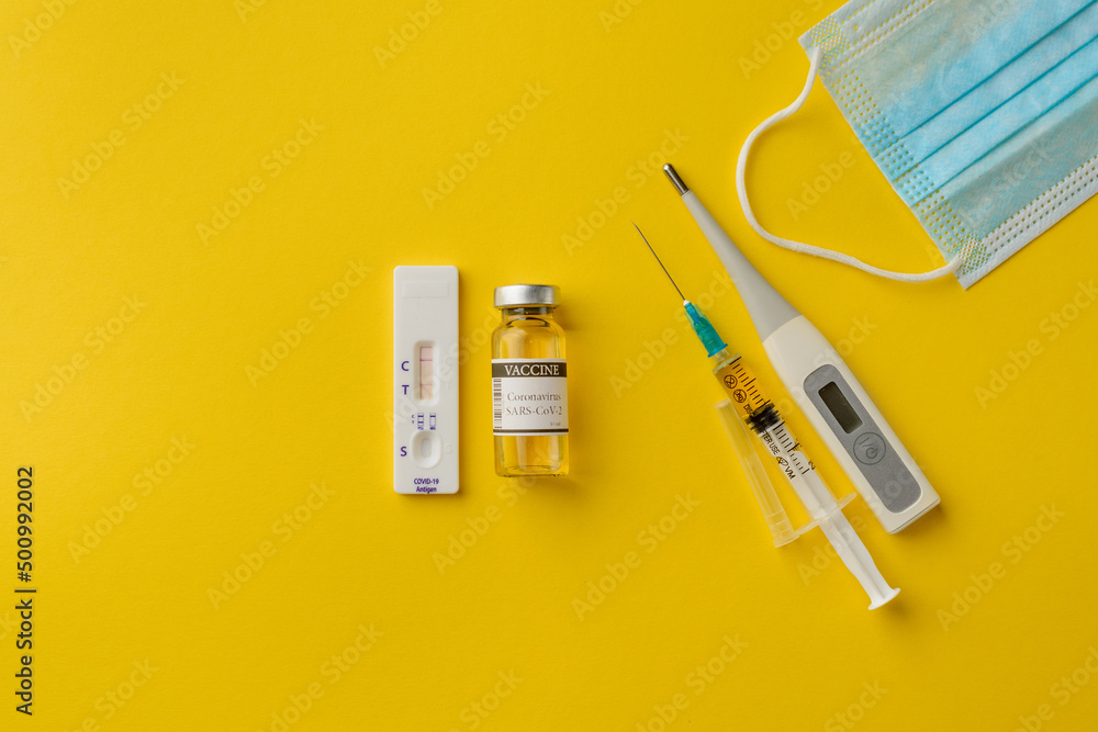 Medical items - a thermometer, a rapid self-diagnosis test for covid-19, an ampoule in a vaccine, a syringe prepared for vaccination, a mask for patients with coronavirus. Yellow background, top view. Medical items - a thermometer, a rapid self-diagnosis test for covid-19, an ampoule in a vaccine, a syringe prepared for vaccination, a mask for patients with coronavirus. Yellow background, top view.