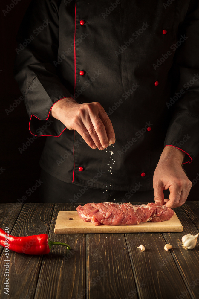 The chef sprinkles raw meat with salt. Preparing beef meat before baking. Working environment in the kitchen.