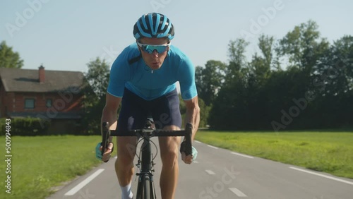 Front view of Caucasian man muscular built, professional racing bicyclist sprint cycling, with posterior raised out of the saddle and pedaling hard, tracking shot.