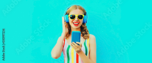 Portrait of happy smiling young woman listening to music in headphones with smartphone on blue background