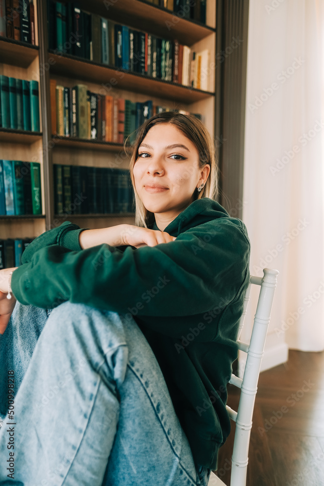 Young caucasian cheerful woman student sitting in a white chair near bookshelf in library. Young caucasian cheerful woman student sitting in a white chair near bookshelf in library.
