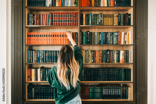 Valokuva Young caucasian woman standing near bookshelf in library and choosing book for reading