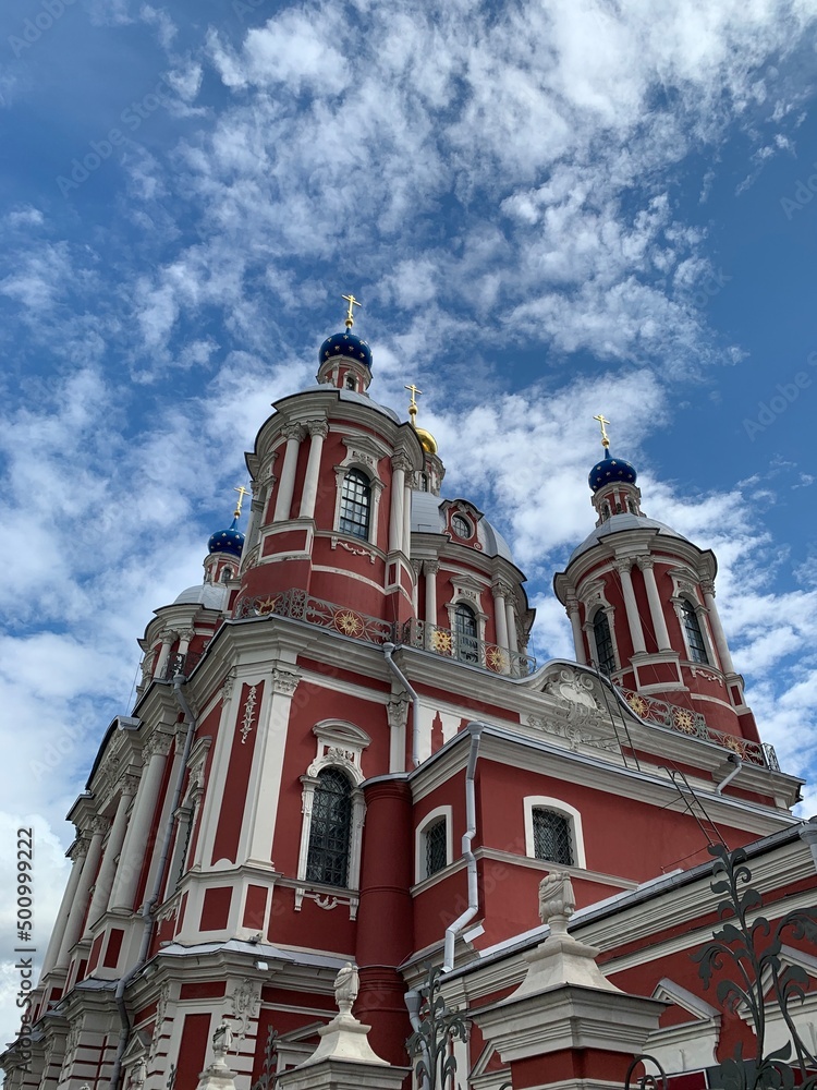 Fototapeta premium Beautiful orthodox church with a chapel in the city with amazing clouds and sky. Bottom view, elongated religious building. The church is red and white with golden domes.