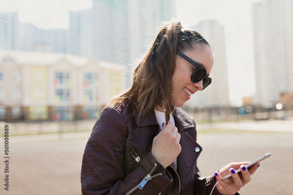 Fototapeta premium Close up portrait of incredible lovely girl with dark hair is looking at smartphone and listening music on city background in sunlight