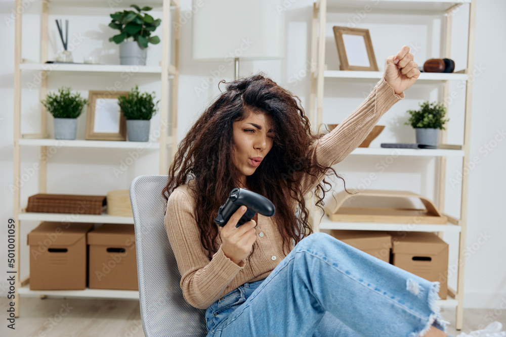 Overjoyed happy lucky tanned curly Latin lady win in game with joystick gamepad sit on chair in home modern interior look aside. Copy space Mockup Banner. Concept game platform