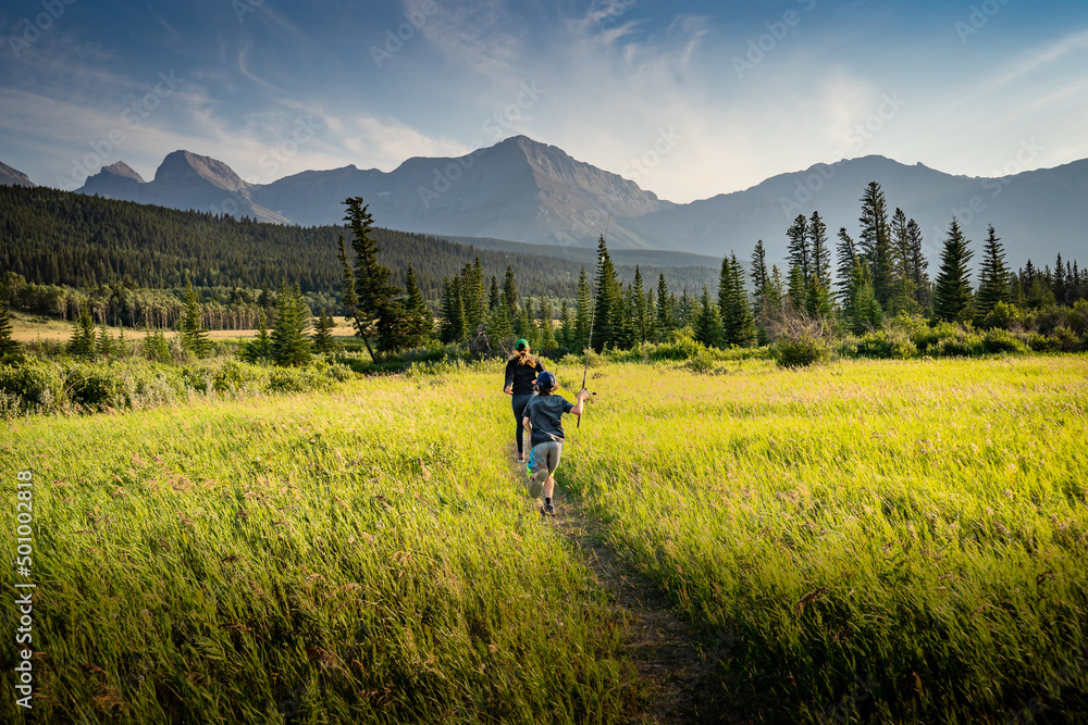 Obraz premium Two children running down a trail across a grassy meadow holding fishing rods in the Canadian Rockies near Crowsnest Pass Alberta Canada during summer.