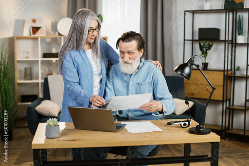 Beautiful mature woman and bearded man in casual outfit using laptop during working together. Pleasant old wife and husband business partners cooperating at bright office or working online from home