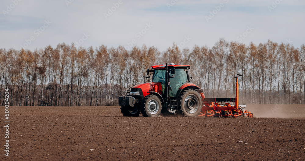 Obraz premium Spring sowing season. Farmer with a tractor sows corn seeds on his field. Planting corn with trailed planter. Farming seeding. The concept of agriculture and agricultural machinery.