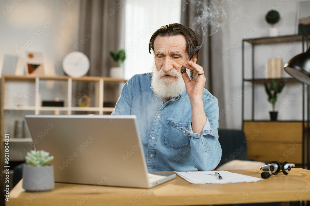 Portrait of handsome elegant mature business man with cigar sitting at desk with laptop during video call and looking at camera. Senior man using modern devices for online conversation at work. Portrait of handsome elegant mature business man with cigar sitting at desk with laptop during video call and looking at camera. Senior man using modern devices for online conversation at work.