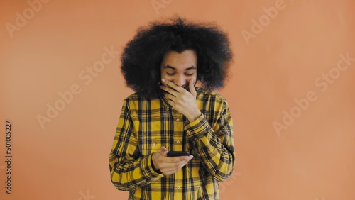 A young man with an African hairstyle on an orange background looks at the phone and is happy. Emotions on a colored background