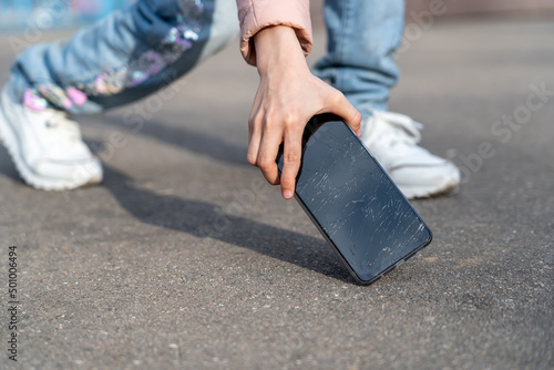 Kid girl picking damaged mobile phone with cracked screen from asphalt outdoors, closeup