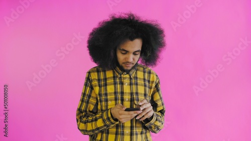 A young man with an African hairstyle on a pink background is talking into his phone.