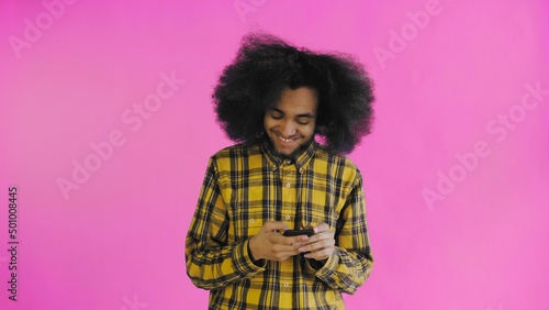 A young man with an African hairstyle on a pink background looks at the phone and happily texts with someone. Emotions on a colored background