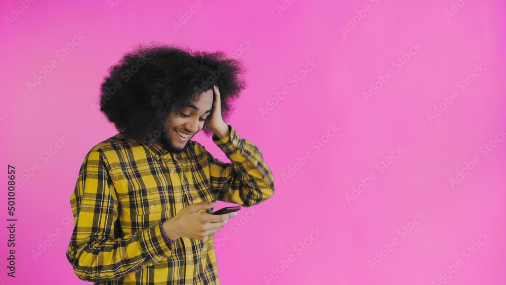 A young man with an African hairstyle on a pink background looks at the phone and is happily surprised. Emotions on a colored background