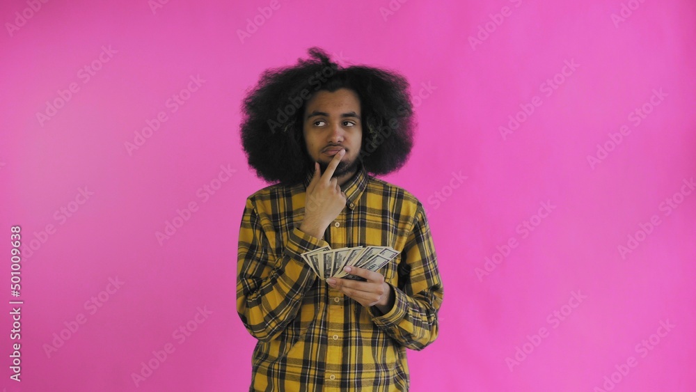 A happy African-American holding money in his hands and thinking about what to spend it on, stands isolated on a pink background A happy African-American holding money in his hands and thinking about what to spend it on, stands isolated on a pink background