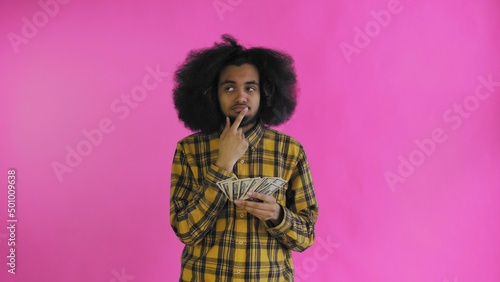 A happy African-American holding money in his hands and thinking about what to spend it on  stands isolated on a pink background