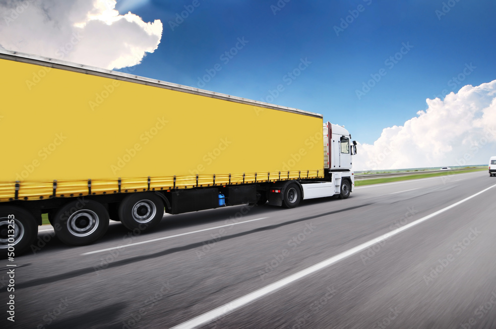 Big truck driving fast with a trailer with space for text on a highway against a sky with clouds