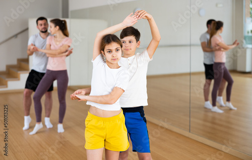 Cute preteen girl and boy siblings enjoying slow foxtrot in pair in dance studio during family training with parents..
