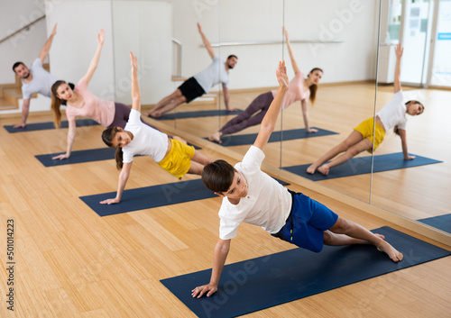 Positive sporty teen boy doing yoga with sister and parents in fitness studio, standing in side plank or Vasisthasana pose..