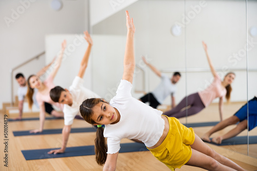 Cute teen girl enjoying yoga class with friendly sporty family in fitness studio, standing in balancing pose Vasisthasana..