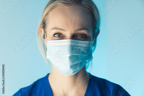 attractive young blonde European nurse smiling under the mask closeup studio shot blue background medical concept. High quality photo