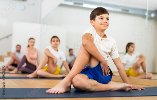 Smiling flexible teen boy sitting on mat in twisting asana Matsyendrasana during yoga class with sportive family in fitness studio