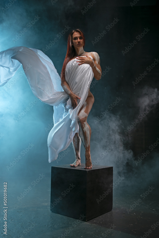 Red-haired girl covers herself with white cloth like an antique statue, posing in the studio