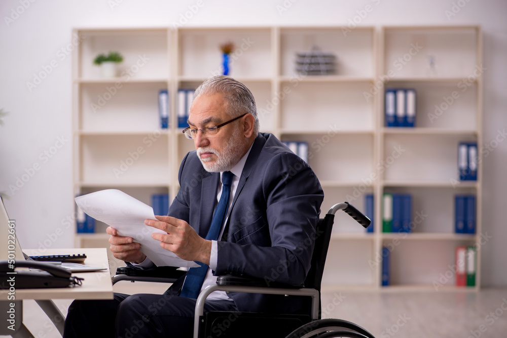 Old male employee in wheel-chair sitting at workplace Old male employee in wheel-chair sitting at workplace