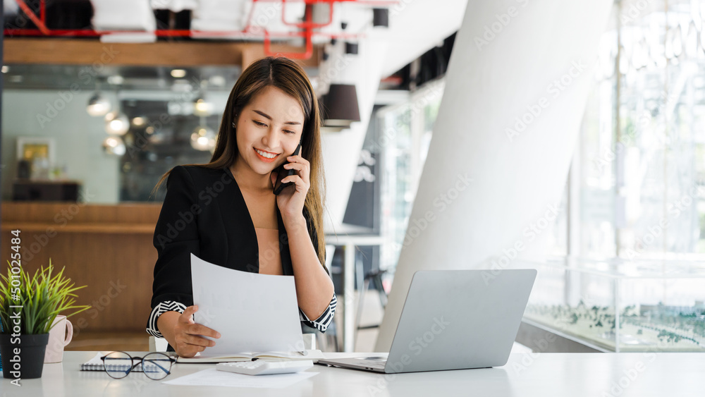 Asian businesswoman talking on mobile phone with customer and looking to work sheet paper in front of laptop computer at office,Office lifestyle concept. Asian businesswoman talking on mobile phone with customer and looking to work sheet paper in front of laptop computer at office,Office lifestyle concept.