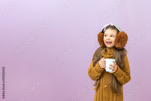 Children is holding a cup with a warm drink in the cold season, a girl in a sweater and warm headphones on an isolated background.