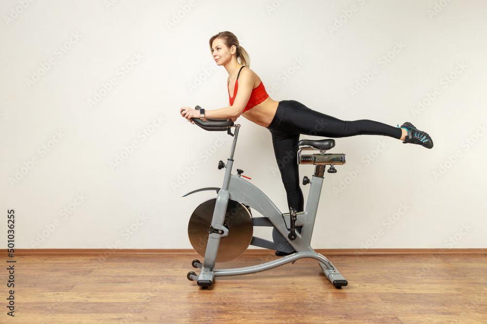 Obraz premium Side view portrait of slim attractive woman standing on exercise bike with raised leg, cardio workout, wearing sports tights and red top. Indoor studio shot on gray wall background.