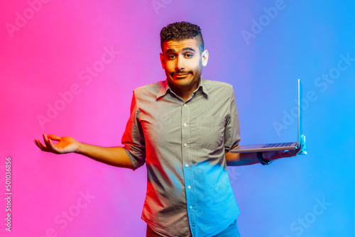 Portrait of puzzled handsome man in shirt standing holding laptop and shrugging, don't know what to do with new project. Indoor studio shot isolated on colorful neon light background. photo