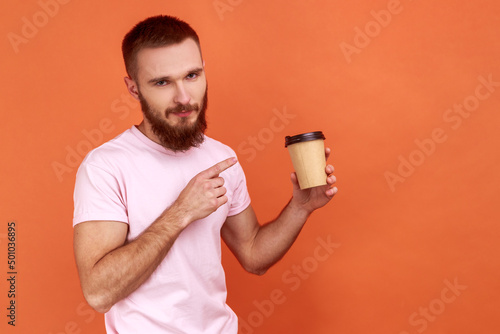 Portrait of handsome bearded man standing holding disposable cup of coffee, pointing at take away beverage and looking at camera, wearing pink T-shirt. Indoor studio shot isolated on orange background