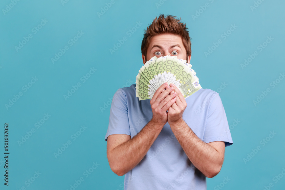 Portrait of handsome young adult bearded man peeping out euro banknotes with playful look, enjoying big jackpot, financial success. Indoor studio shot isolated on blue background. - obrazy, fototapety, plakaty 
