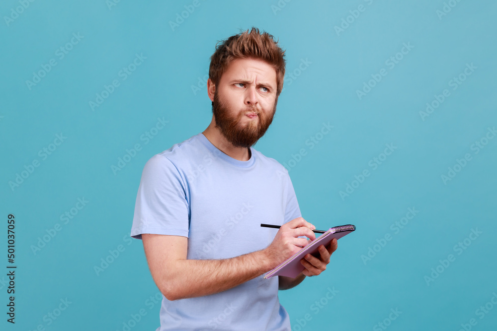 Portrait of bearded man making notes in paper notebook, writing business idea, future plans, looking away with thoughtful expression. Indoor studio shot isolated on blue background. Portrait of bearded man making notes in paper notebook, writing business idea, future plans, looking away with thoughtful expression. Indoor studio shot isolated on blue background.